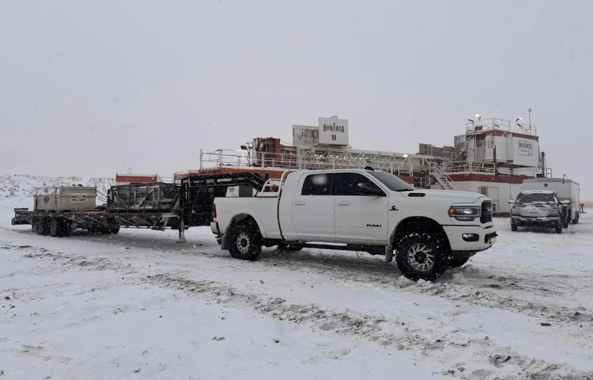 Rig Dawgs truck pulling a loaded gooseneck trailer on a snowy lease, drilling rig in the background